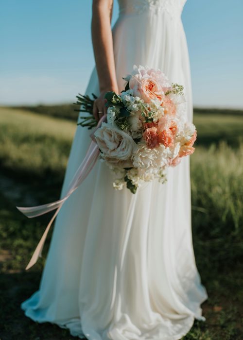 A bride in a flowing white gown holds a colorful bouquet in a sunny field.