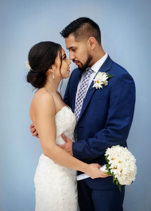 Bride and groom sharing a tender moment with bouquet against a blue background.