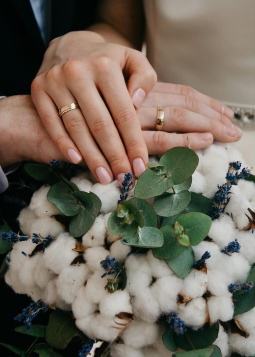 Close-up of couple's hands with gold wedding bands over a floral bouquet.